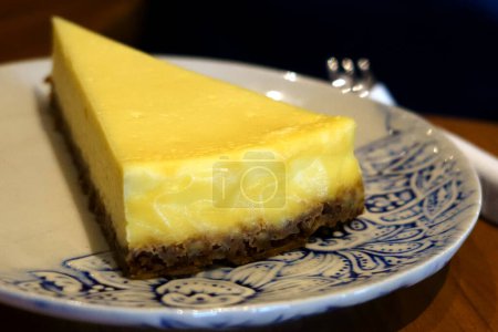 Foto de Una tentadora rebanada de tarta de queso se sienta en un plato azul y blanco con dibujos. Un tenedor descansa a su lado, listo para cavar. - Imagen libre de derechos