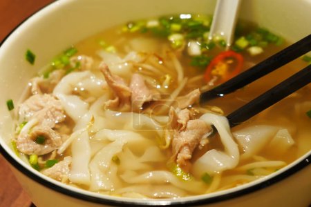 A close-up photo of a steaming bowl of pho. The broth is light brown and there are thin slices of beef, white onions, and green onions floating on top. There are also white chopsticks on the side of the bowl.