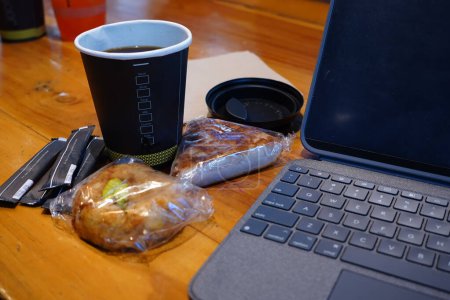 a workplace setup featuring a cup of coffee, a slice of bread, and an iPad propped up on a wooden table.