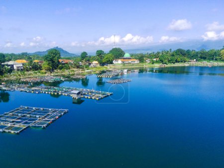 Téléchargez les photos : Vue aérienne d'une petite ville au bord du lac avec une mosquée, entourée d'arbres et de maisons. Des cages à poissons sont visibles dans l'eau près de Ranu ou du lac Klakah, Lumajang, Java oriental, Indonésie. - en image libre de droit