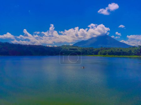 Téléchargez les photos : Une vue aérienne d'un lac bleu clair avec un spectaculaire, nuages plafonnés montagne en arrière-plan. Des arbres sempervirents bordent le rivage près de Ranu ou du lac Klakah, Lumajang, Java oriental, Indonésie. - en image libre de droit