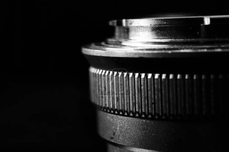 La imagen es un primer plano en blanco y negro de una lente de cámara. La lente tiene varios elementos de vidrio curvado y una carcasa metálica.