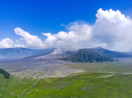 Luftaufnahme des Vulkans Bromo, Ostjava, Indonesien mit Rauch, der aus dem Krater aufsteigt. Der Vulkan ist von einem grünen Feld und einem blauen Himmel umgeben.