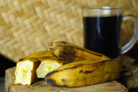 un plátano al vapor a la mitad y un plátano entero en una tabla de cortar de madera junto a una taza de café negro.