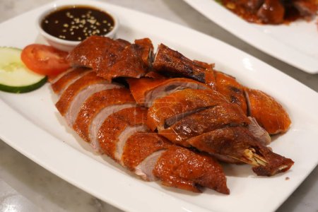 Ein Nahaufnahme-Foto einer gebratenen Ente mit knuspriger Haut auf einem weißen Teller. Serviert wird es mit dunkler Soße und geschnittenen Zwiebeln.