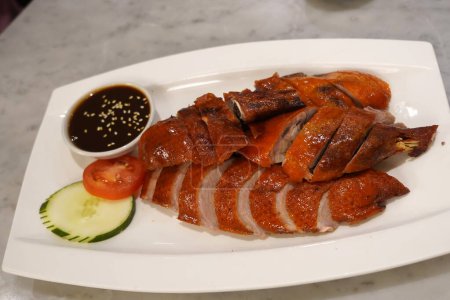 A close up photo of a roasted duck with crispy skin on a white plate. It is served with a dark sauce and sliced scallions.