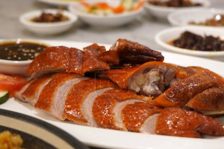 A close up photo of a roasted duck with crispy skin on a white plate. It is served with a dark sauce and sliced scallions.