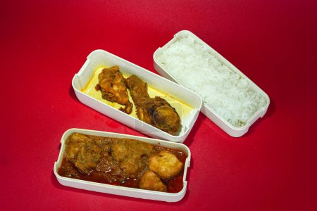 A lunchbox with compartments containing chicken, rice, and curry sits on a red table.