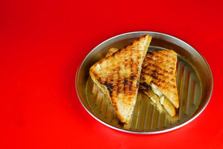 Téléchargez les photos : Un sandwich au fromage grillé brun doré sur une plaque rectangulaire en métal. La plaque est sur une table rouge avec une surface lisse. - en image libre de droit