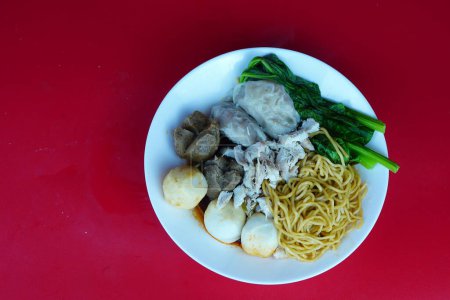 A round white bowl filled with chicken noodles, vegetables, and meatballs in a brown sauce. Isolated