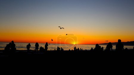 Téléchargez les photos : Coucher de soleil suggestif sur la plage avec des silhouettes de personnes regardant le coucher du soleil - en image libre de droit