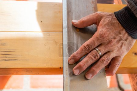 Téléchargez les photos : Homme plus âgé travaillant à la maison avec une scie à main. Latino couper le bois et réparer. Outils de menuiserie. Main avec anneau au premier plan. Espace de copie. - en image libre de droit