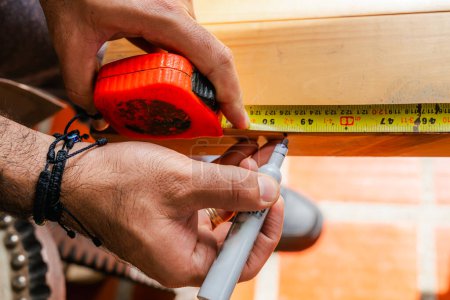 Téléchargez les photos : Homme mesurant le bois avec flexomètre et marquage au crayon travaillant dans sa maison. Latino dans son atelier. - en image libre de droit