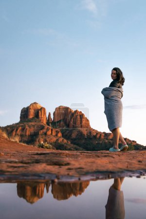 Téléchargez les photos : Young good looking female woman standing with blanket wrapped around her looking at camera with red rock cathedral rock butte lit up by incredible sunset colors during blue hour in Sedona Arizona USA. - en image libre de droit