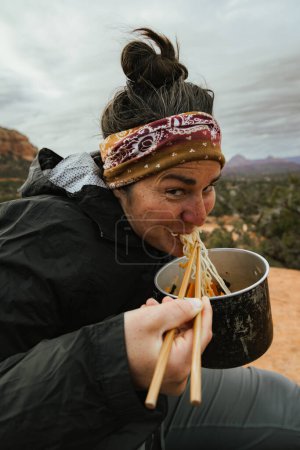 Foto de Young happy beautiful caucasian woman in her 30s smiles at camera eating ramen out of camping pot with chopsticks in Sedona Arizona USA - Imagen libre de derechos