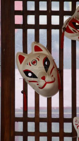 Traditional Japanese Kitsune Mask Hanging on Wooden Lattice with Light Filtering Through, Featuring Red and White Design Elements in Cultural Setting