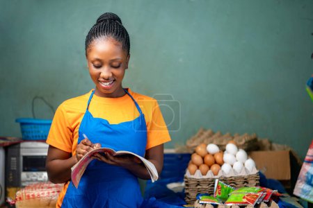 Foto de Hermosa dama africana con un libro, productos alimenticios la rodearon - tendero haciendo balance - Imagen libre de derechos