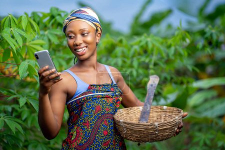 Foto de Señora africana, con teléfono inteligente y cesta en una granja-mujer negra surfeando en las redes sociales-concepto agrícola - Imagen libre de derechos