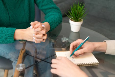 Téléchargez les photos : Gros plan des mains d'une femme avec un cahier et une plante d'intérieur sur la table.Deux femmes sont assises à une table en verre, l'une prend des notes dans un cahier. Concentration sélective - en image libre de droit
