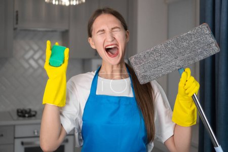 Photo for Young woman in a uniform apron holds a mop and sponge in her hands. She is screaming, unhappy, tired and very depressed. She hates her job cleaning the apartment. Cleaning. Women 's work - Royalty Free Image