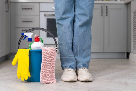 Woman in uniform stands next to a blue bucket filled with chemicals, cleaning products, a rag against the background of the kitchen. Cleaning company concept, house cleaning