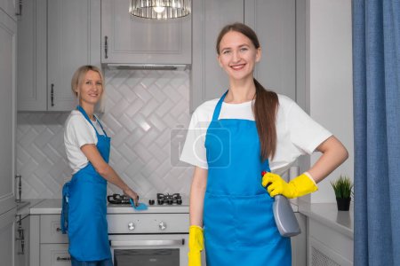 Beautiful young woman in an apron with detergent and a sponge looks at the camera, in the blurry background, a young female assistant is cleaning the kitchen. Cleaning company concept, cleaning team