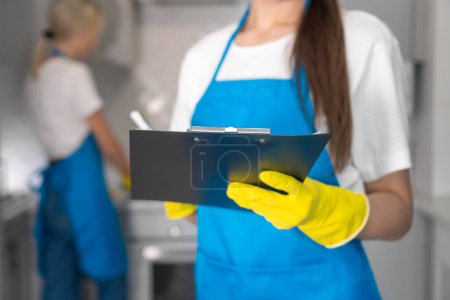 Close-up of a young cleaning woman in an apron writes down a report on the work done on a paper tablet against the background of a blurred employee. Cleaning company in the kitchen, teamwork
