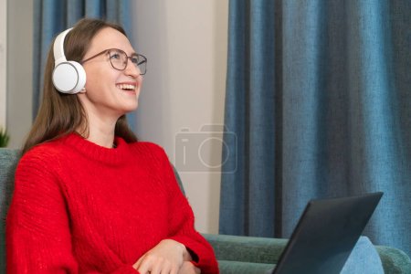 Lächelnde junge Frau mit Kopfhörer, Brille und rotem Pullover sitzt auf dem Sofa, den Kopf vor Lachen zurückgeworfen, einen Laptop auf dem Schoß. Konzept positiver Emotionen