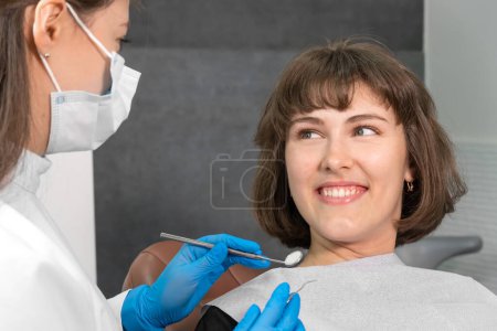 Photo for Young smiling woman is sitting in a dental chair and looking at a dentist who is holding dental instruments in his hands to check the oral cavity - Royalty Free Image