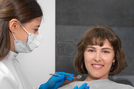 Téléchargez les photos : Une jeune femme souriante est assise sur une chaise dentaire et regardant dans la caméra, le dentiste tient des instruments dentaires pour examiner la cavité buccale - en image libre de droit