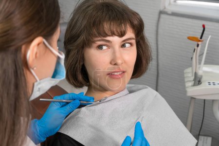 Photo for Young smiling woman is sitting in a dental chair and looking at a dentist who is holding dental instruments in his hands to check the oral cavity - Royalty Free Image