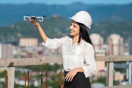 Foto de Hermosa ingeniera geodésica sonriente en un casco blanco sostiene un dron a lo largo del brazo en un sitio de construcción. Ingeniero Arquitectónico e Inspector de Seguridad - Imagen libre de derechos