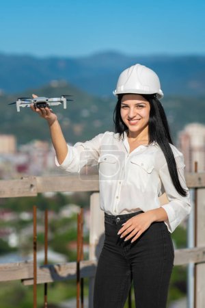 Foto de Una hermosa ingeniera geodésica sonriente con un casco blanco sostiene un dron a lo largo de un brazo en un sitio de construcción y mira a la cámara. Ingeniero de arquitectura e inspector de seguridad.Foto vertical - Imagen libre de derechos