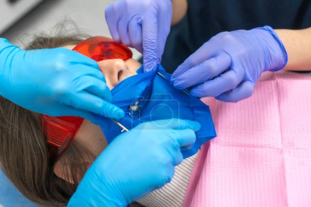 Photo for Process of installing a dental cofferdam for dental treatment on the jaw of a young woman wearing protective glasses in dentistry. Dentist uses a dental dam to isolate the tooth - Royalty Free Image