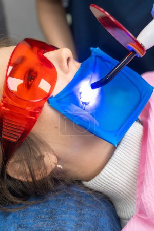 Photo for Close-up of a beautiful young woman in protective glasses in a dentist's chair with her mouth open at the time of tooth polymerization. Work of a polymerization lamp in the hands of a dentist - Royalty Free Image