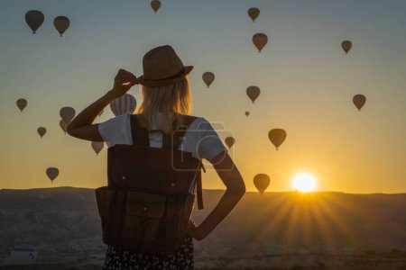 Eine Touristin mit Hut und Rucksack steht allein an der Spitze und bewundert den Ballonflug während eines wunderschönen Sonnenuntergangs, Kappadokien, Türkei. Fabelhaftes Kappadokien mit einem Ballonflug im Morgengrauen..