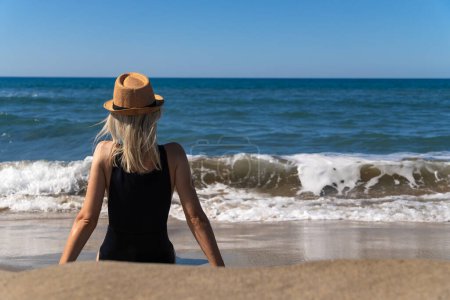 Jeune femme mince en maillot de bain noir avec un chapeau est assis sur une plage de sable en face de la mer avec des vagues par une journée ensoleillée, vue arrière. Jeune fille pense et se détendre au bord de la mer.