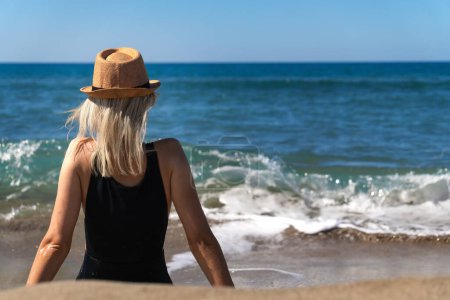 Slender young woman in a black swimsuit with a hat is sitting on a sandy beach opposite the sea with waves on a sunny day, rear view. Young girl is thinking and relaxing by the sea.