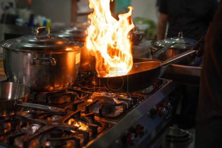 Les mains du chef tiennent un wok avec le feu sur une cuisinière à gaz. Gros plan sur les mains du chef cuisinier cuisinant de la nourriture sur le feu. Chef prépare la nourriture dans une cuisine professionnelle.