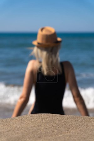 Gros plan d'un endroit vide sur le sable sur un fond flou d'une jeune femme en maillot de bain noir avec un chapeau sur la plage par une journée ensoleillée.La jeune fille réfléchit et se relaxe au bord de la mer.Photo verticale