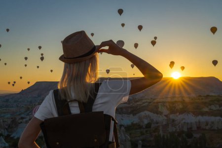 Female tourist in a hat with a backpack stands alone at the top and admires the flight of balloons during a beautiful sunset, Cappadocia, Turkey.Fabulous Cappadocia with a flight of balloons at dawn.