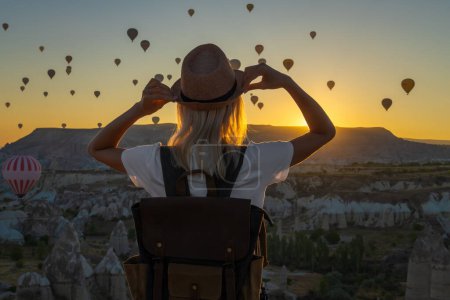 Eine Touristin mit Hut und Rucksack steht allein an der Spitze und bewundert den Ballonflug während eines wunderschönen Sonnenuntergangs, Kappadokien, Türkei. Fabelhaftes Kappadokien mit einem Ballonflug im Morgengrauen..