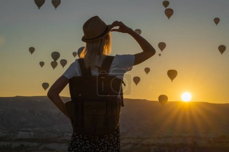 Junge Touristin mit Hut und Rucksack bewundert die atemberaubende Landschaft der Heißluftballonflüge über dem Tal der Liebe im Moment eines wunderschönen Sonnenaufgangs, Kappadokien, Türkei.