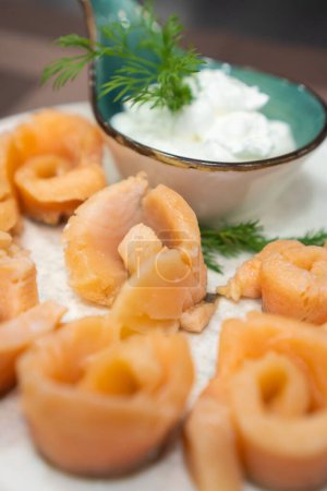 Close-up of pickled salmon pieces on a white plate with sauce and dill on the table. Beautiful serving of pickled salmon. Vertical photo
