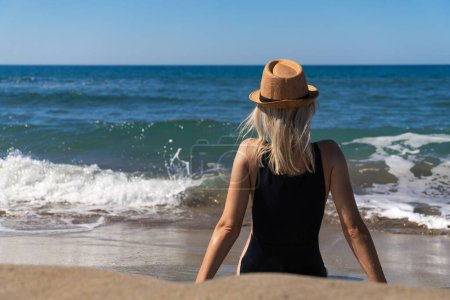 Schlanke junge Frau in schwarzem Badeanzug und Hut sitzt an einem sonnigen Tag an einem Sandstrand gegenüber dem Meer mit Wellen, Rückansicht. Junges Mädchen denkt und entspannt am Meer.