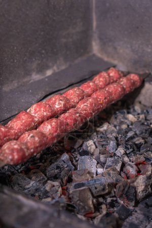 Nahaufnahme eines rohen Döners mit Gewürzen auf Spießen wird auf Kohlen mit Rauch gekocht, selektiver Fokus. Vertikales Foto