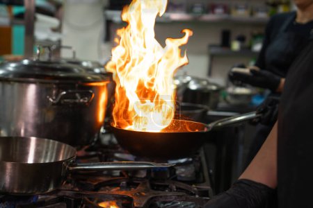 Téléchargez les photos : Les mains du chef tiennent un wok avec le feu sur une cuisinière à gaz. Gros plan sur les mains du chef cuisinier cuisinant de la nourriture sur le feu. Chef prépare la nourriture dans une cuisine professionnelle. - en image libre de droit