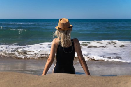 Mujer joven delgada en un traje de baño negro con un sombrero está sentado en una playa de arena frente al mar con olas en un día soleado, vista trasera. Chica joven está pensando y relajándose junto al mar.