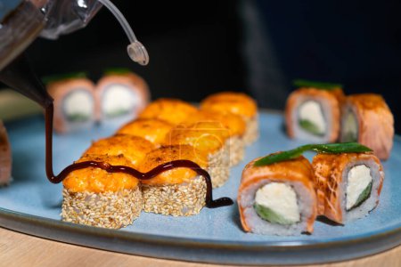 Zbliżenie szefa kuchni wylewając sos teriyaki na pieczone bułki sushi na talerzu w kuchni w restauracji.