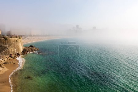 Dichter Nebel an einem sonnigen Tag. Atlantik, Strand, Ufer und Stadtgebäude im Nebel. La Coruna, Galicien, Spanien.